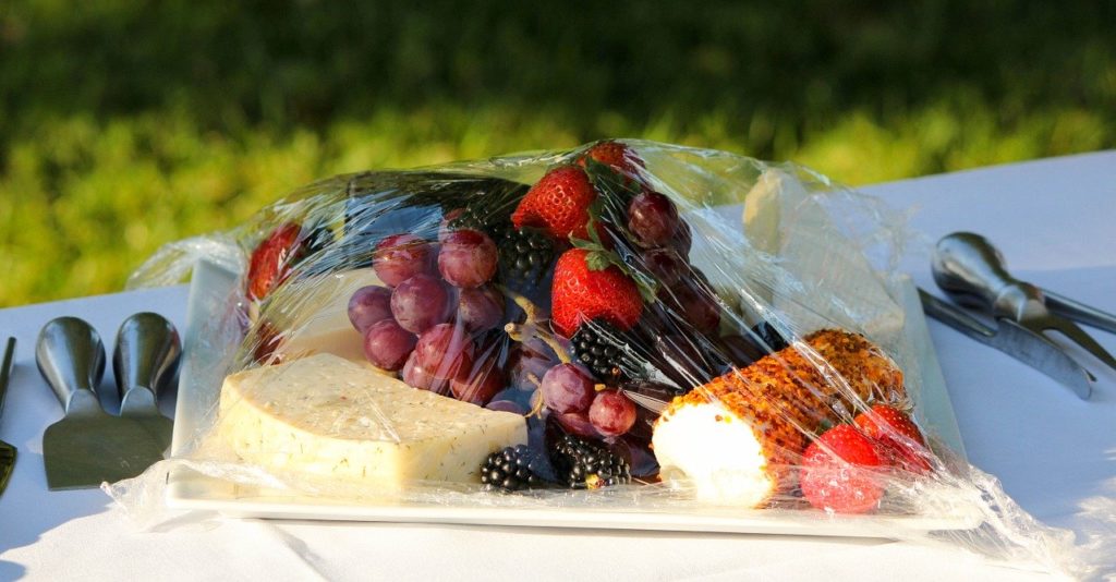 Queijo, frutas e pão embalados com celofane sobre uma tolha em cima da grama